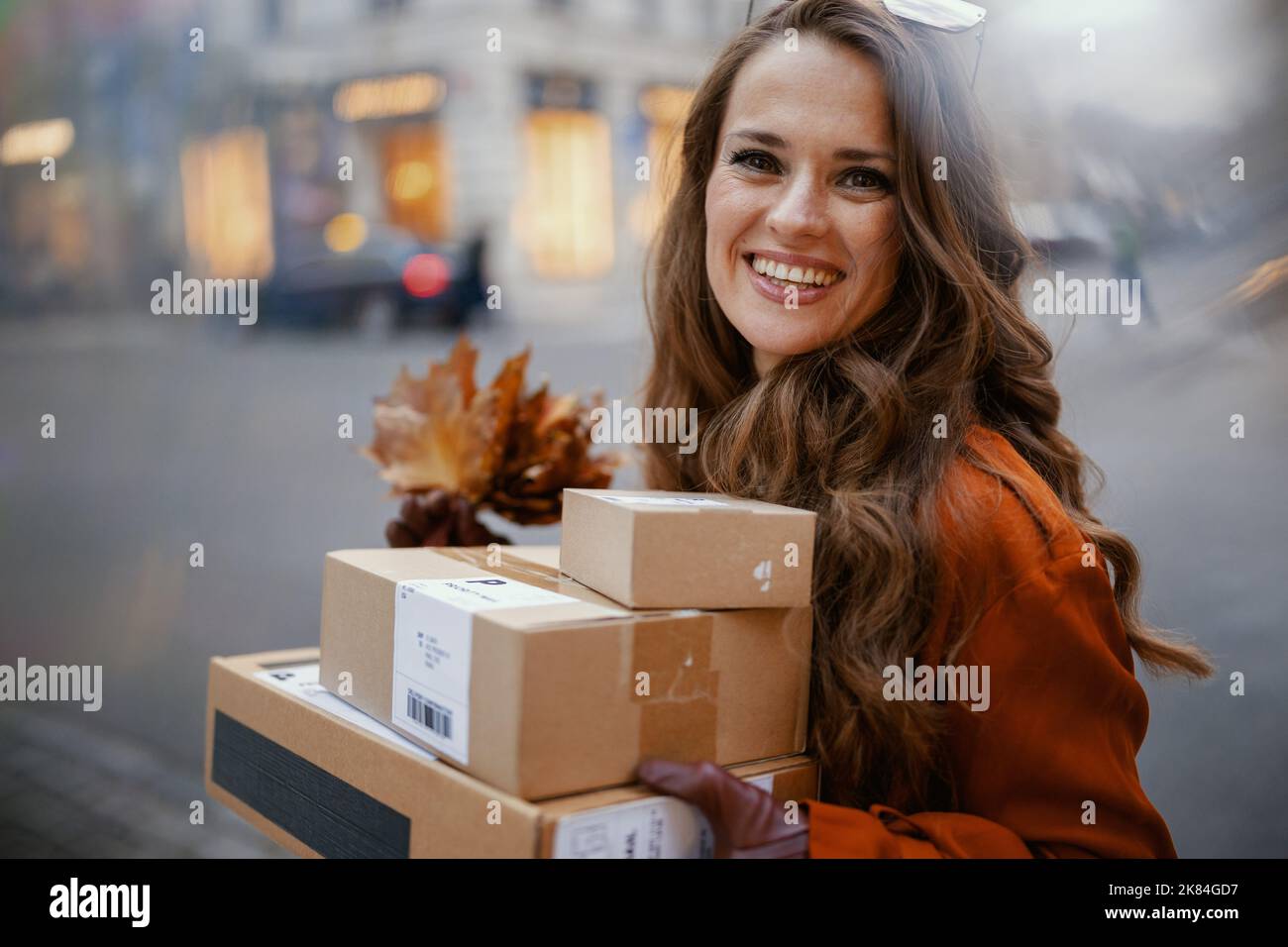 Ciao settembre. Felice donna trendy in trench arancione con pacchi e foglie gialle autunnali in città. Foto Stock