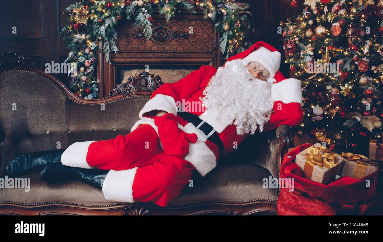 Babbo Natale dorme in una stanza decorata vicino all'albero di Natale con un sacco di regali, l'uomo in costume è sdraiato sul divano. Vacanza e presenta concetto. Foto Stock