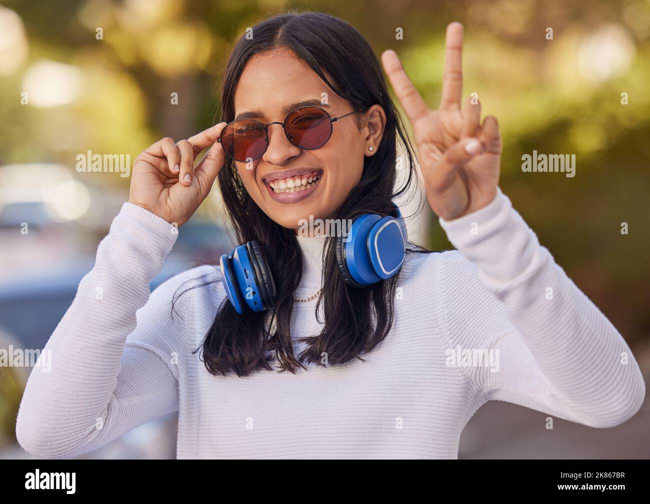 Musica, cuffie e segno di pace mano di una donna dal messico in natura su una passeggiata. Ritratto di una persona con un sorriso felice, occhiali da sole e gesto Foto Stock