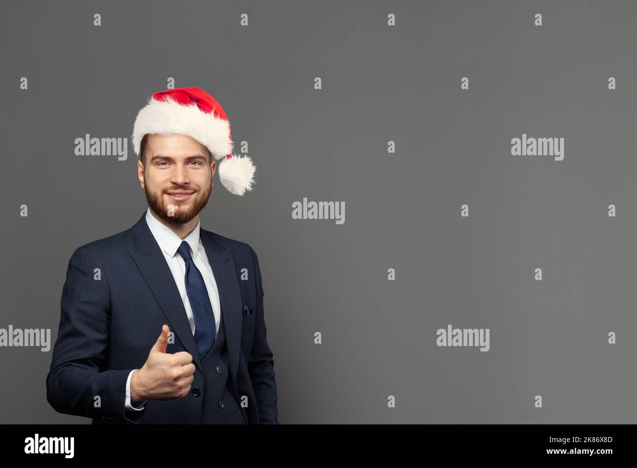 Gioioso uomo d'affari nel cappello di Natale sta mostrando pollice in su e guardando sullo sfondo grigio della bandiera della parete dello studio Foto Stock