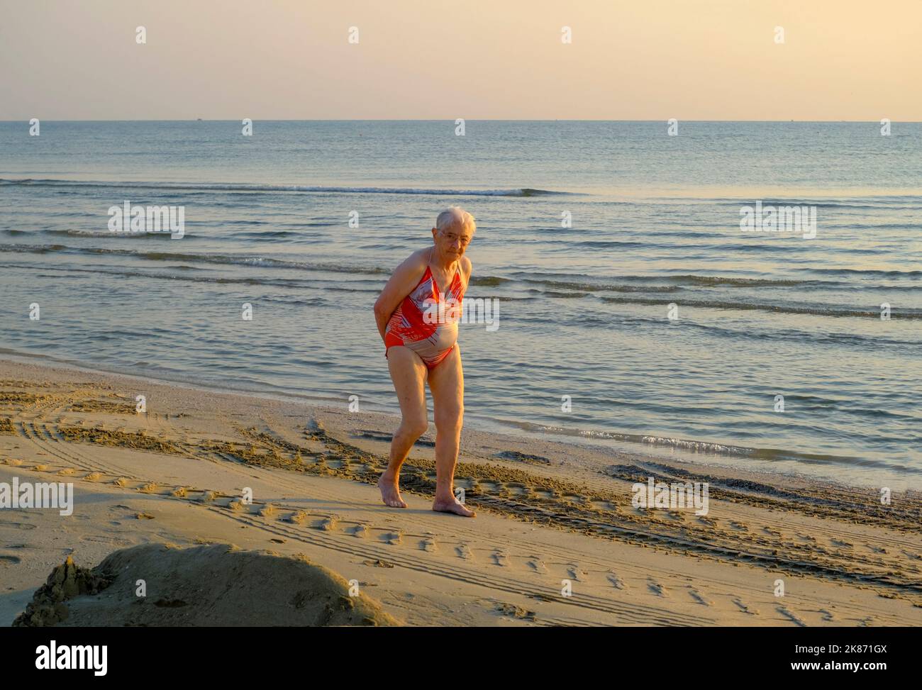 Luglio 2022 Senigallia, Italia: Donna anziana che cammina sulla spiaggia attraverso il mare tramonto e l'orizzonte. Foto Stock