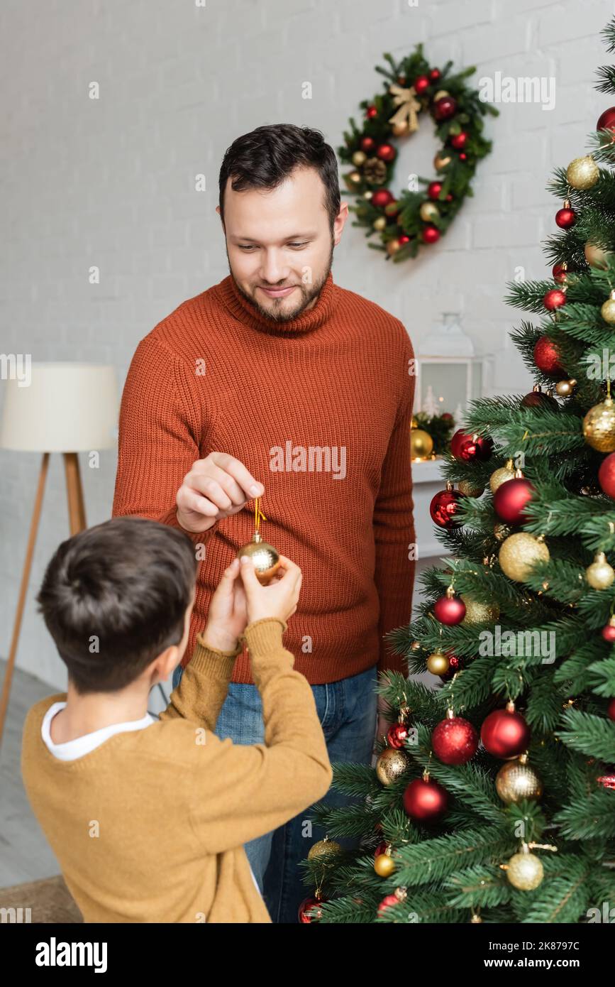 papà sorridente che dà la sfera di natale al figlio che decora l'abete verde a casa Foto Stock