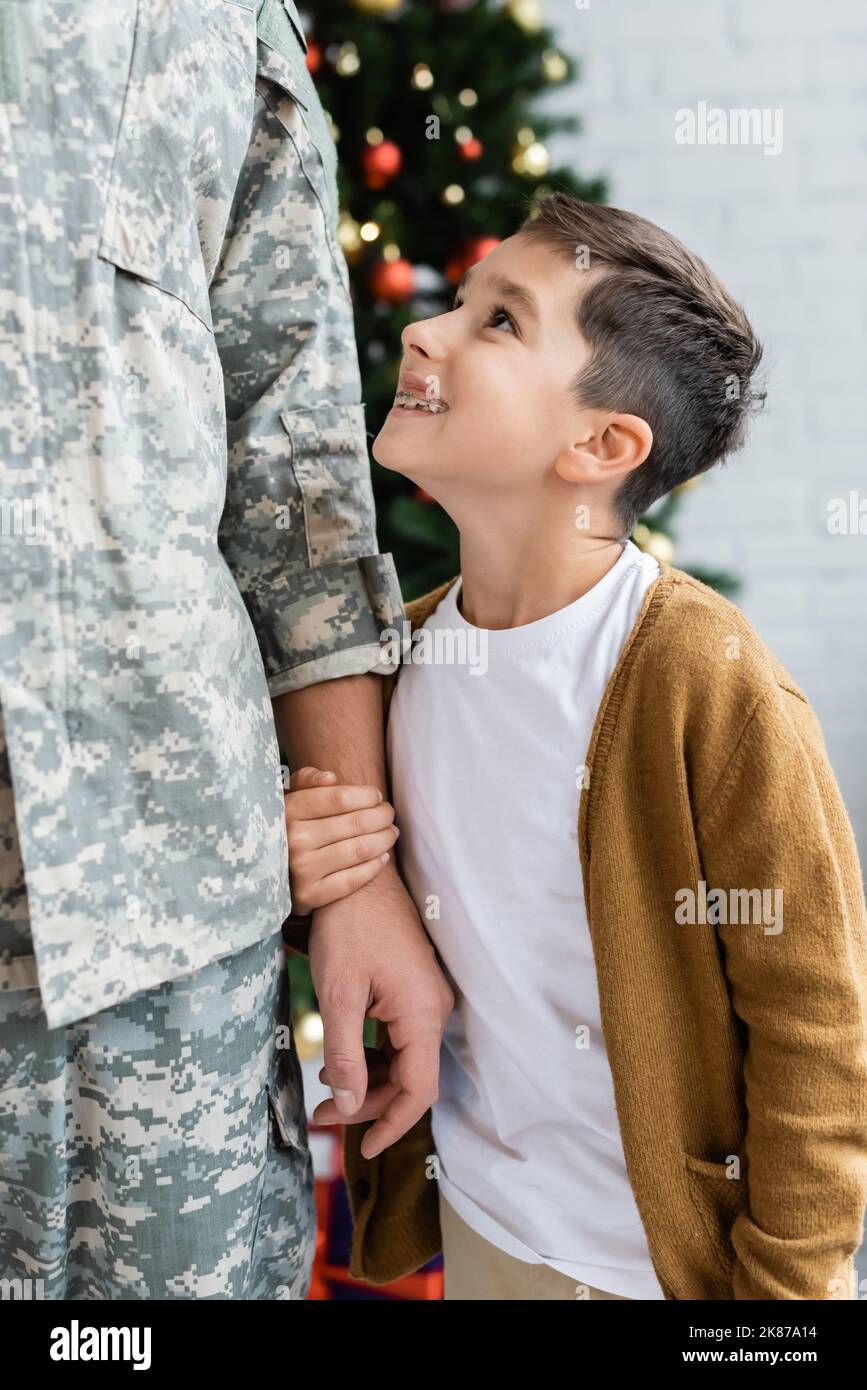 ragazzo compiaciuto guardando il padre in uniforme militare e tenendo la mano a casa Foto Stock
