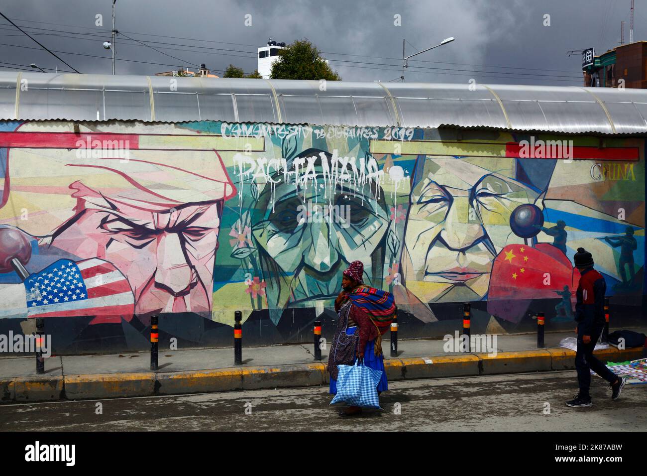 Signora Aymara che cammina davanti al murale mostrando Pachamama (Madre Terra) come una donna anziana triste fiancheggiata da Donald Trump e Xi Jinping, la Ceja, El Alto, Bolivia Foto Stock