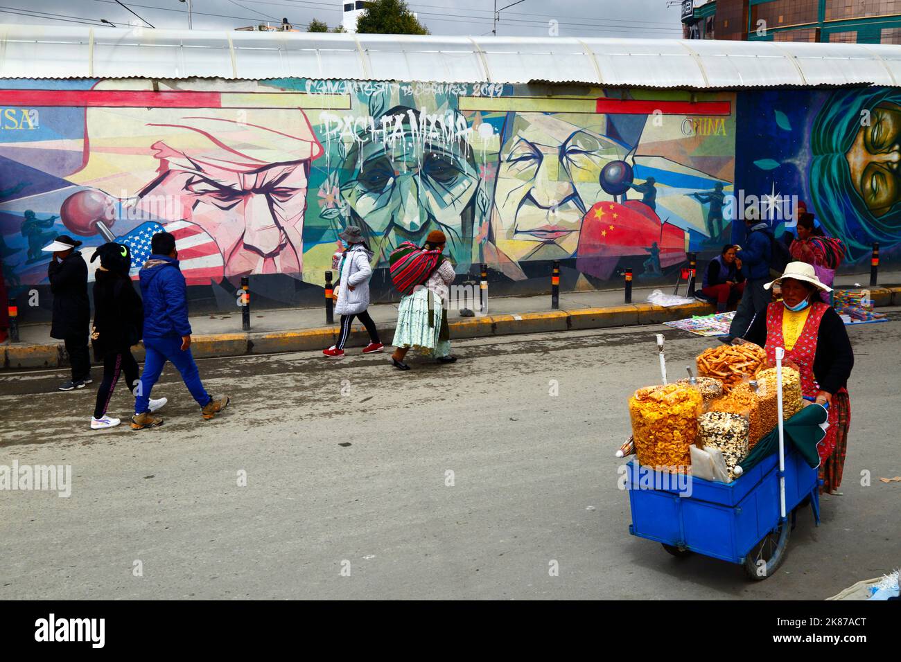 Il venditore ambulante che cammina il murale che mostra Pachamama (la madre terra) come donna anziana triste fiancheggiata da Donald Trump e Xi Jinping, la Ceja, El Alto, Bolivia Foto Stock