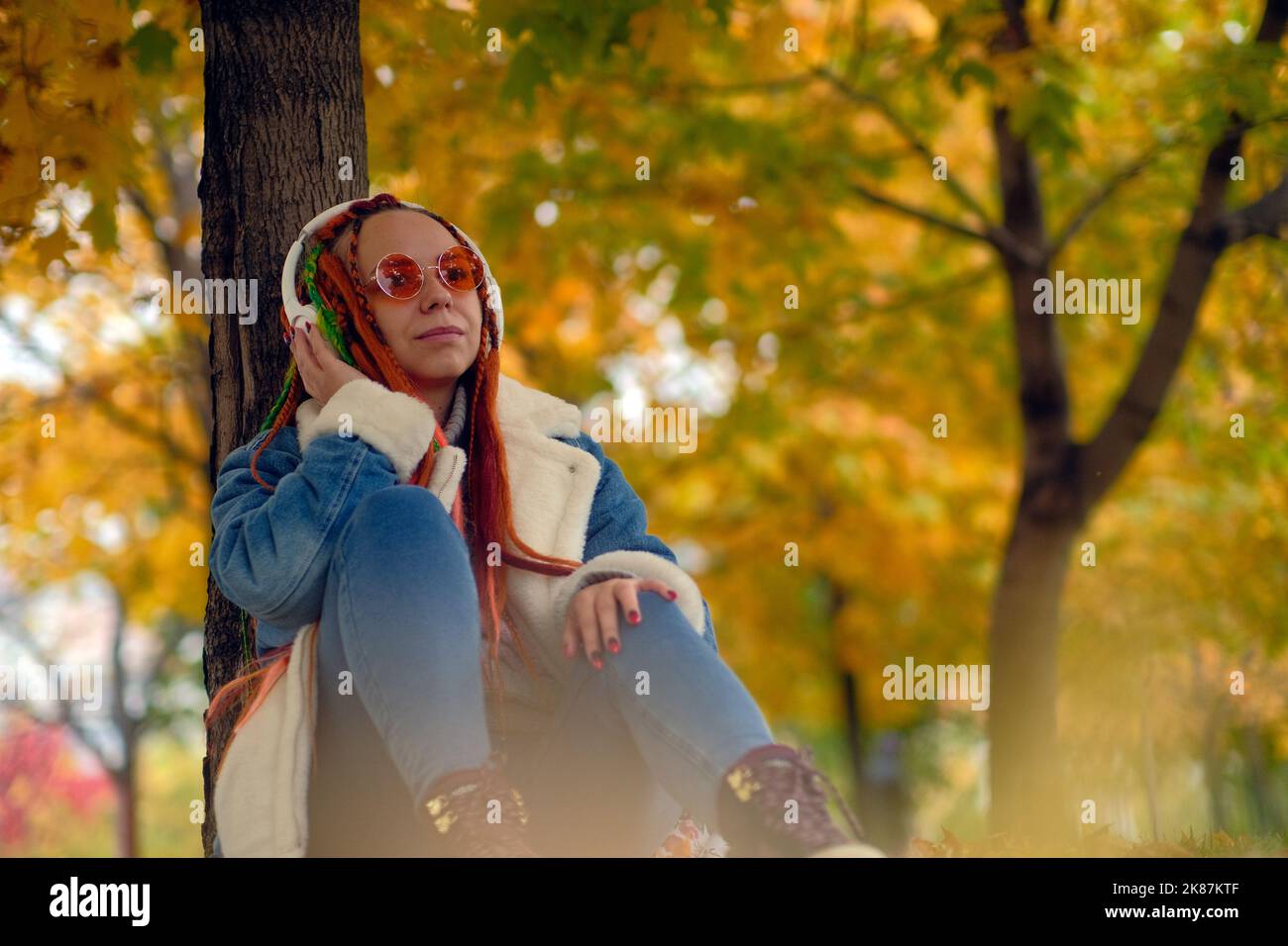 Signora con i greggi che ascoltano la musica nel parco. Giovane donna con i variopinti greggi in cuffie wireless che ascoltano la musica mentre si appoggia sull'albero Foto Stock