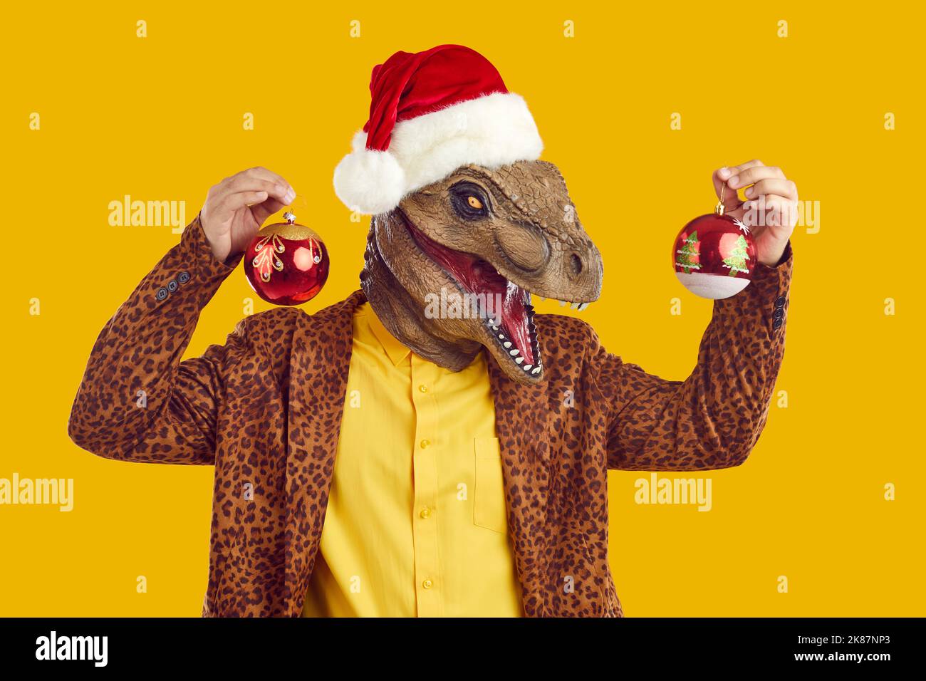 Uomo divertente in maschera di dinosauro, cappello di Natale e tuta leopardo che tiene palle di albero di Natale Foto Stock
