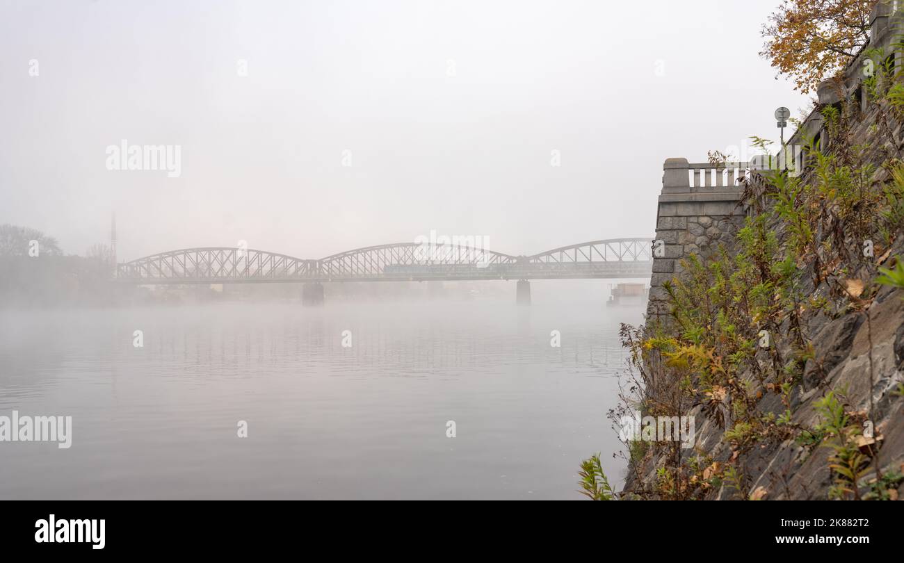 Argine del fiume Moldava a Praga in una mattina d'autunno misteriosa. Gli archi del ponte ferroviario di Vyšehradský sono visibili nella nebbia, con un treno che passa. Foto Stock