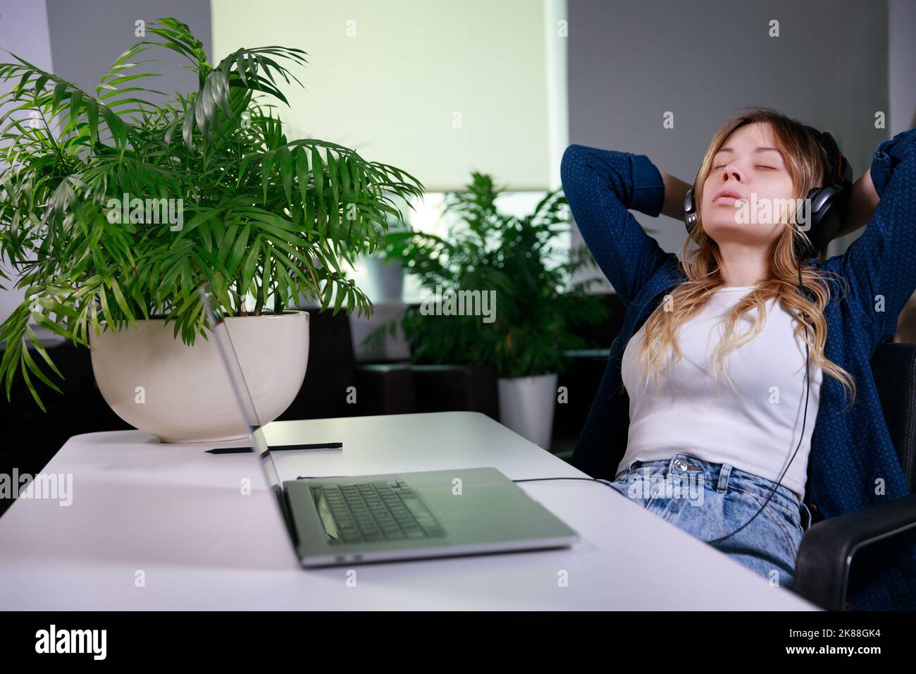 la donna in ufficio si siede e si appoggia su una sedia e ascolta con le cuffie Foto Stock