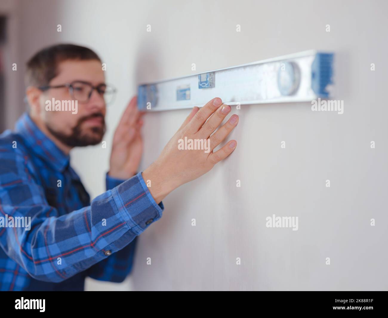 Giovane uomo in tuta di lavoro blu che fa appartamento di riparazione. Concetto di ristrutturazione della casa. Tiro corto di bel giovane uomo bearded che tiene attrezzo di livello durante la sede Foto Stock