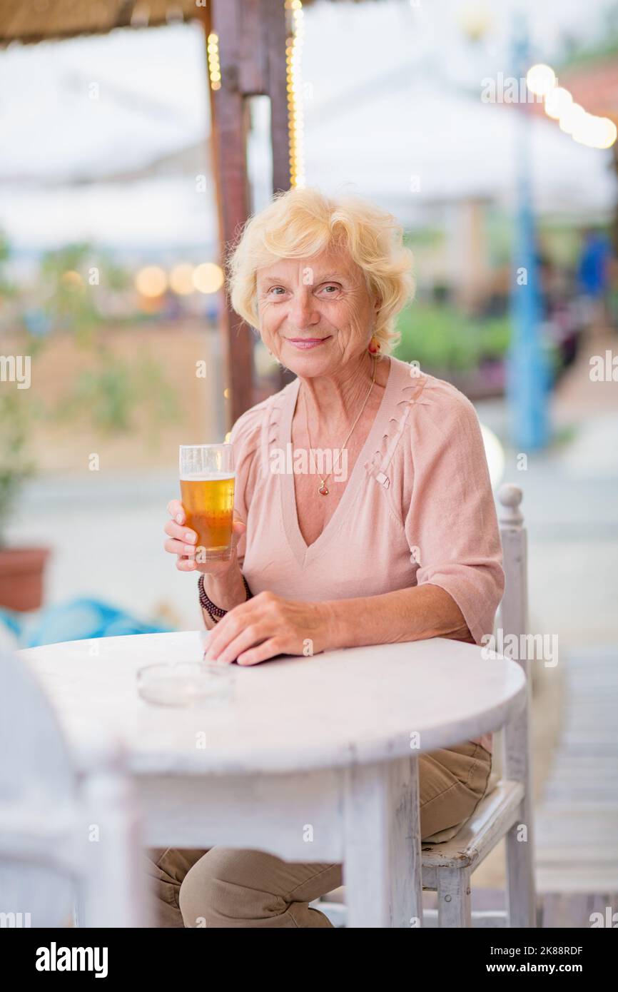 Donna matura seduta a un tavolo in un bar estivo e bere birra Foto Stock