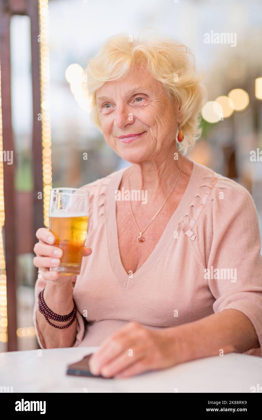 Donna matura seduta a un tavolo in un bar estivo e bere birra Foto Stock
