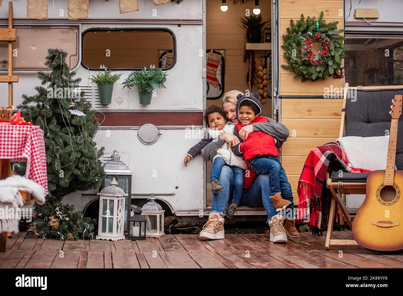 La madre caucasica gioca divertendosi con i bambini afroamericani sulla casa del rimorchio della porta d'ingresso. Giovane donna abbraccia bacia figlio, figlia. L'elogio di famiglia Foto Stock