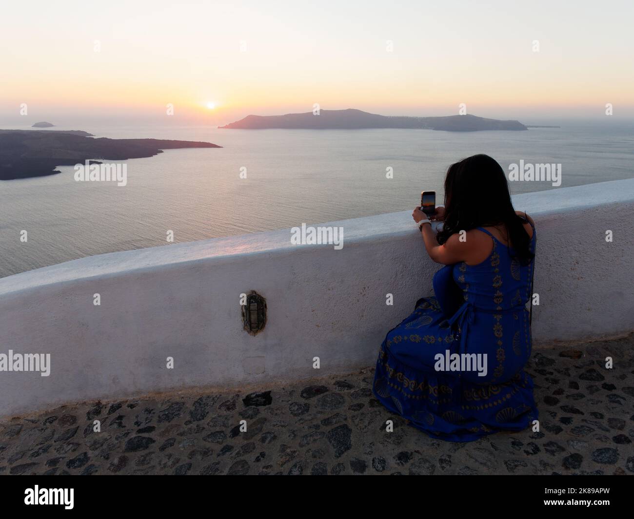 Lady cattura il tramonto sulla Thirasia sul suo telefono da un punto di vista a Fira. Greco Egeo Cicladi Isola di Santorini. Foto Stock