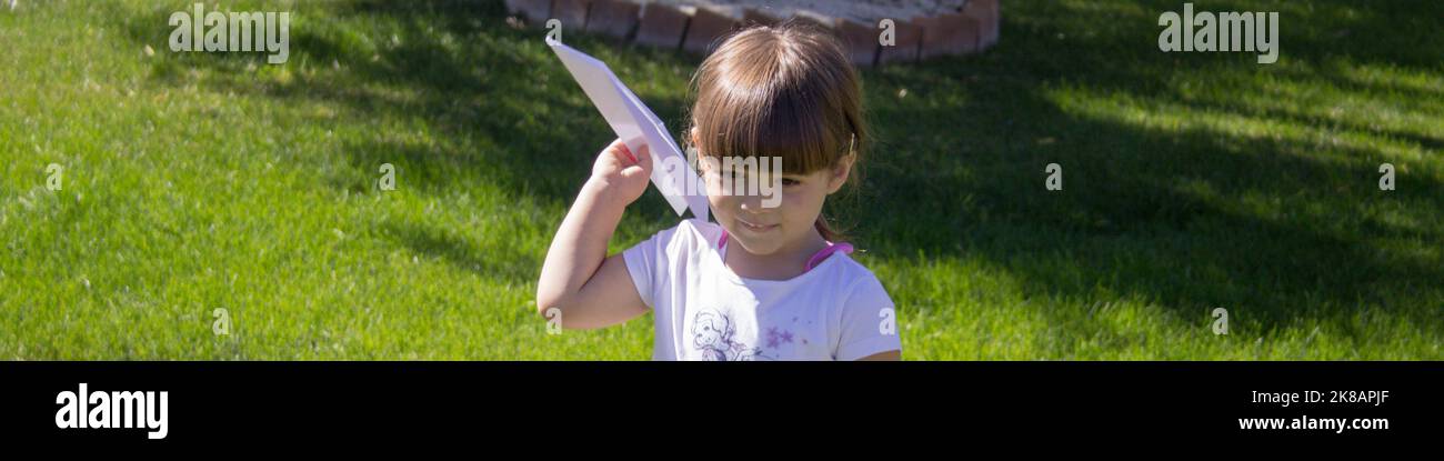 Adorabile bambina che gioca a volare un aereo di carta nel giardino a casa. Giochi all'aperto. Banner orizzontale Foto Stock