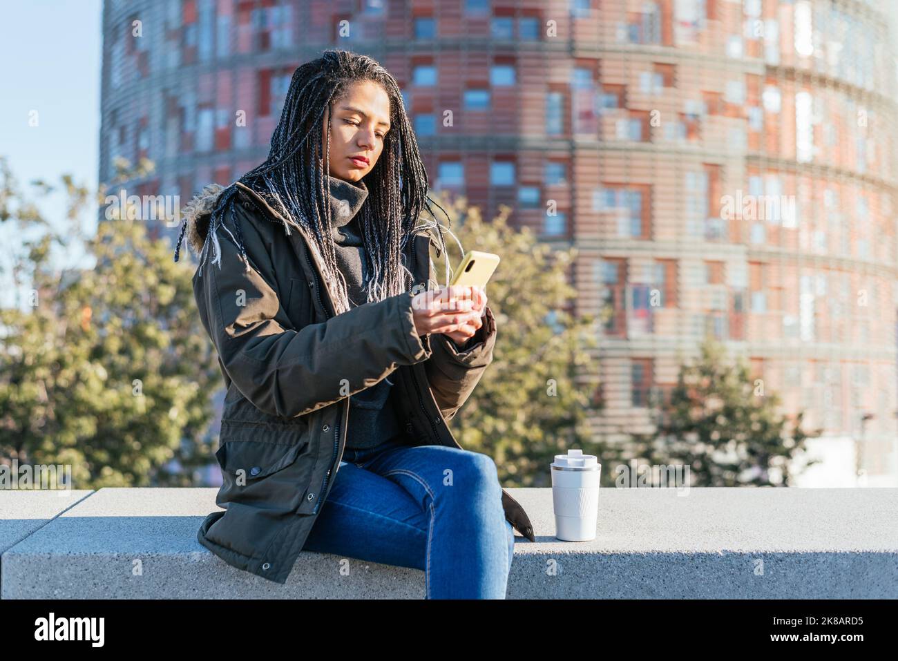 Donna ispanica focalizzata con dreadlocks messaggi di testo sul cellulare mentre si siede sulla panca con thermos di caffè caldo in città Foto Stock