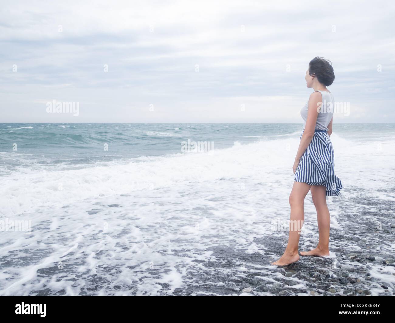 Una donna felice saluta il mare tempestoso sulla spiaggia di ciottoli. Donna con i capelli ruffled con il vento. Concetto di Wanderlust. Vacanza sulla costa del mare. Foto Stock