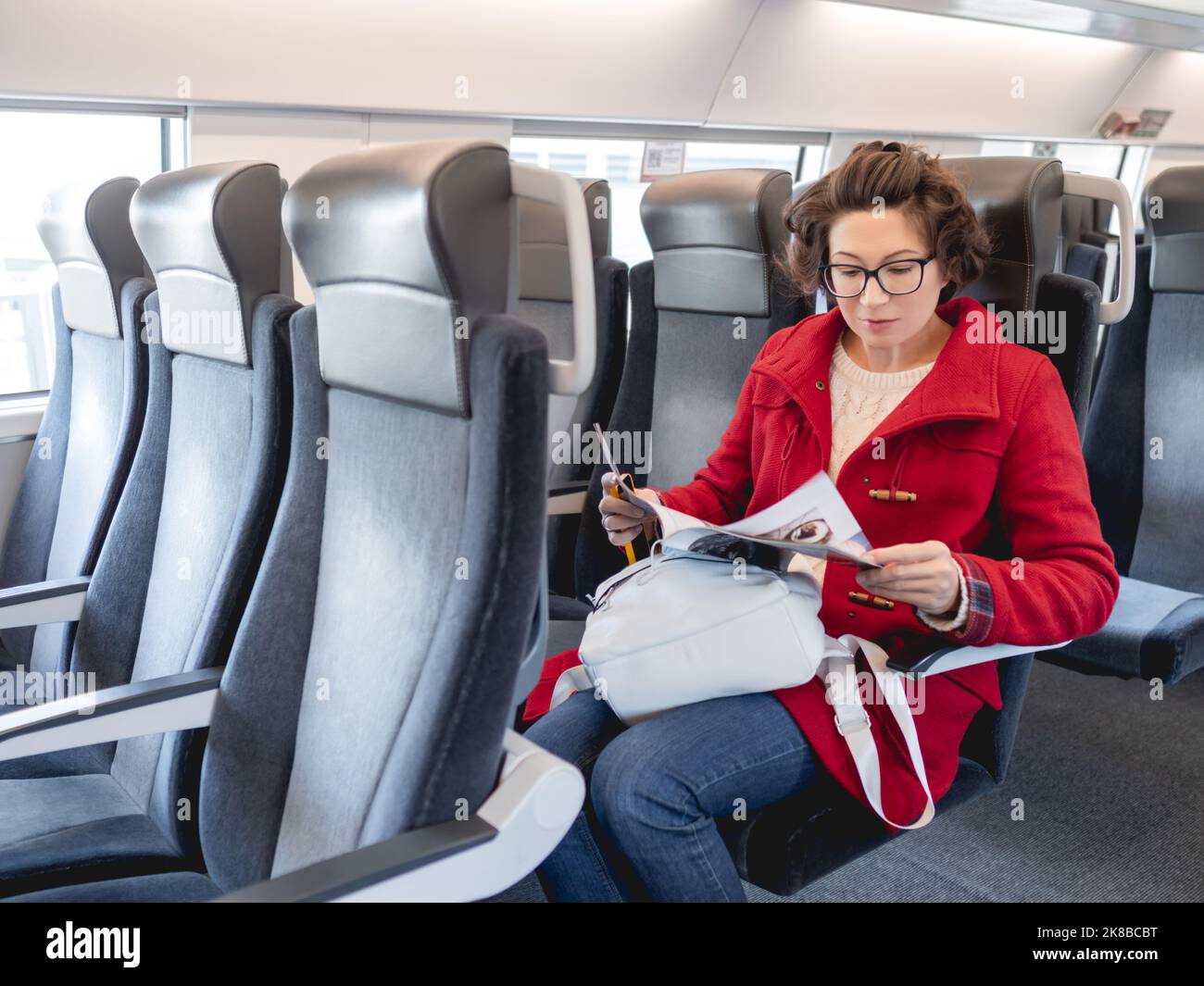 Donna in dufflé rosso legge la rivista in treno suburbano. Informazioni sui supporti stampati. Viaggiare con un veicolo terrestre. Foto Stock