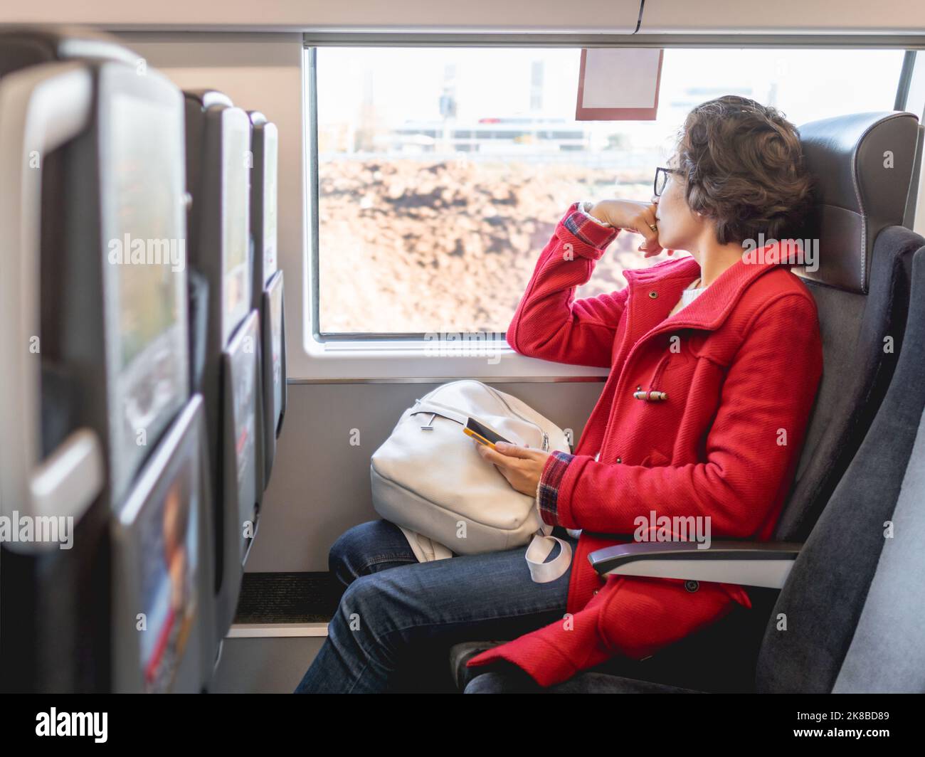 Donna sorridente in dufflé rosso con su smartphone si siede vicino alla finestra del treno suburbano. Viaggiare con un veicolo terrestre. Foto Stock