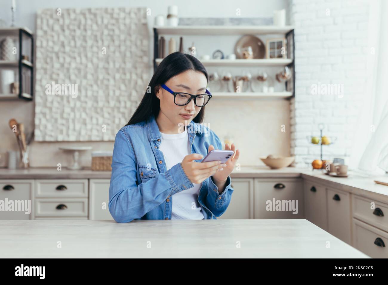 Giovane bella donna asiatica seduta in cucina a casa e utilizzando il telefono cellulare. Scorre il feed delle notizie, si trova sui social network, controlla la posta. Foto Stock