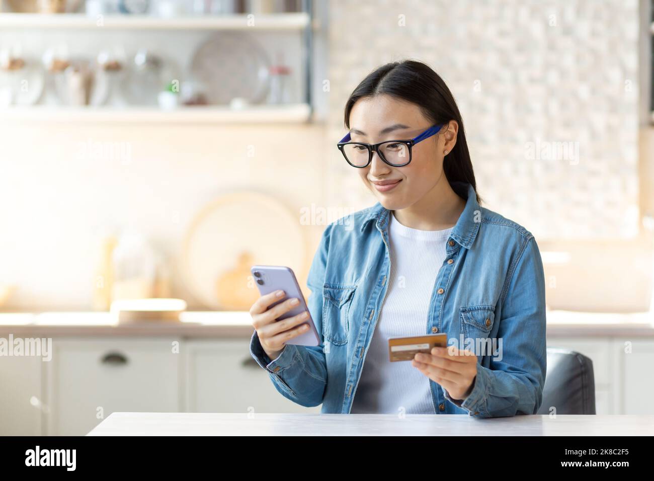 Shopping online. Una giovane sorridente bella donna asiatica fa un ordine di shopping online e paga con un conto senza contanti. È in possesso di un telefono e di una carta di credito. Seduti a casa in cucina. Foto Stock