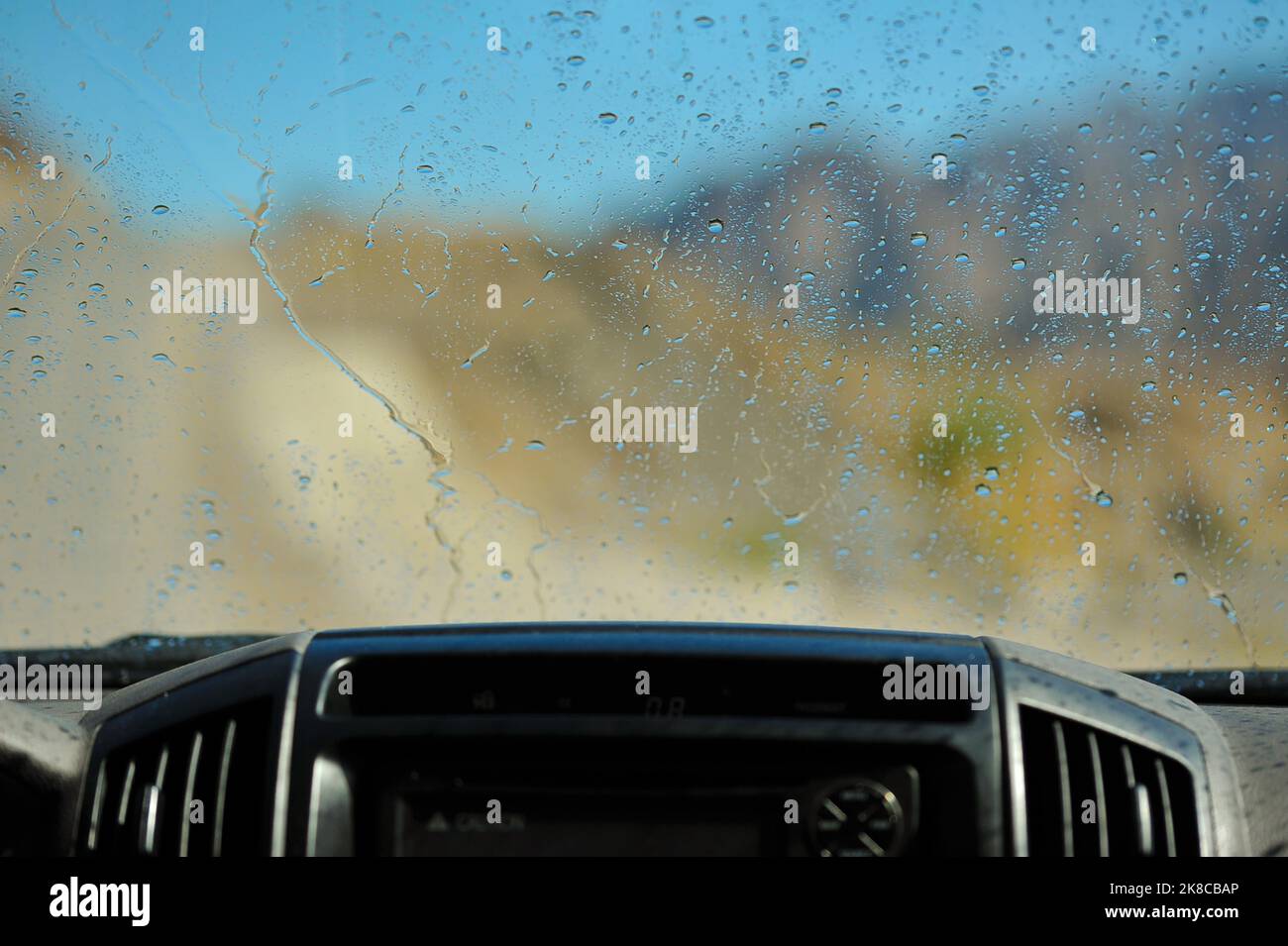 Primo piano del parabrezza con gocce d'acqua dall'interno di un'auto. Vista sfocata della strada, delle montagne e del cielo blu in una giornata estiva, parte superiore del cruscotto davanti. Foto Stock