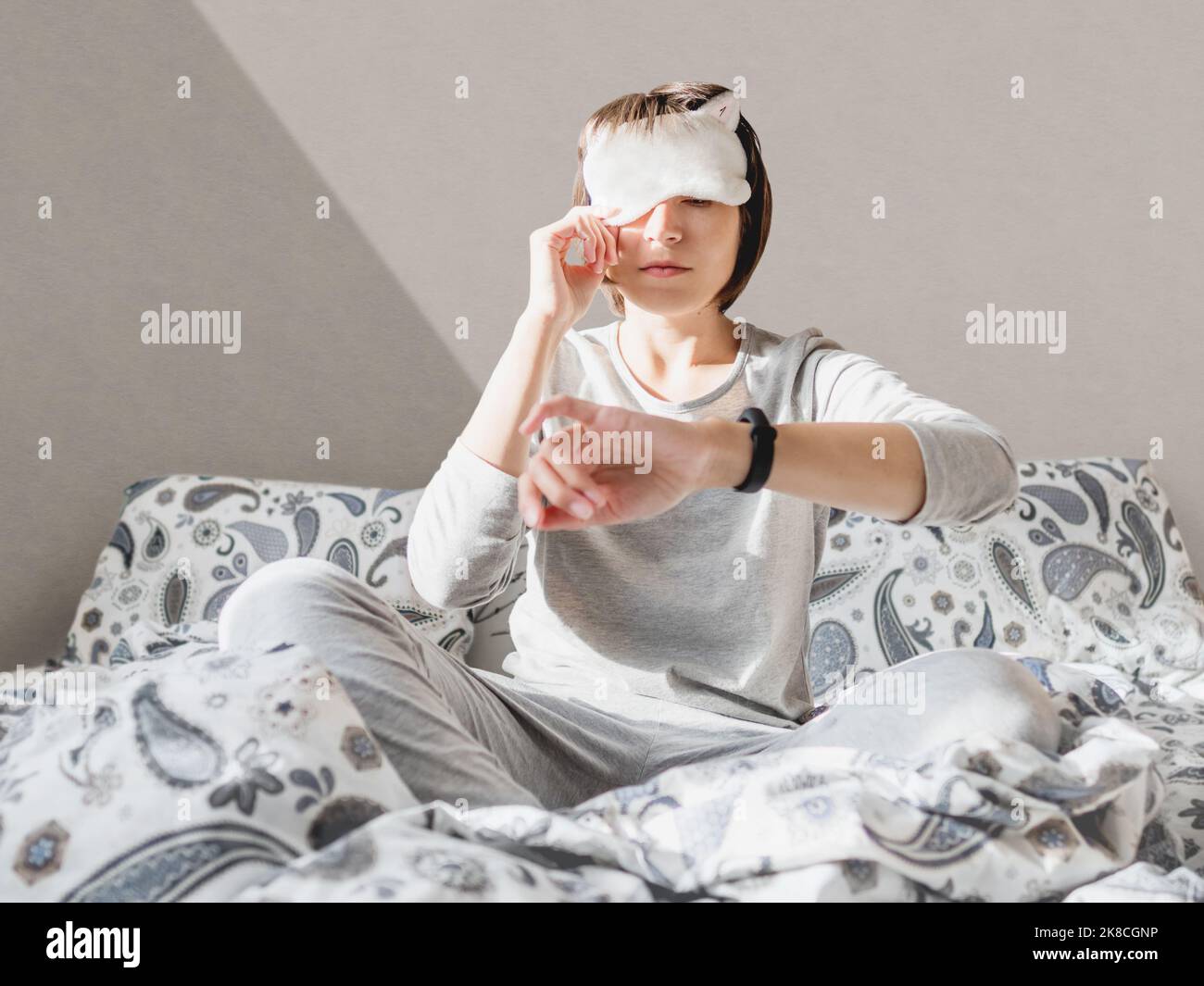 Donna addormentata in pajama grigia e maschera addormentata sul viso. Si è appena svegliata e si siede a letto. Mattina presto in casa accogliente. Foto Stock