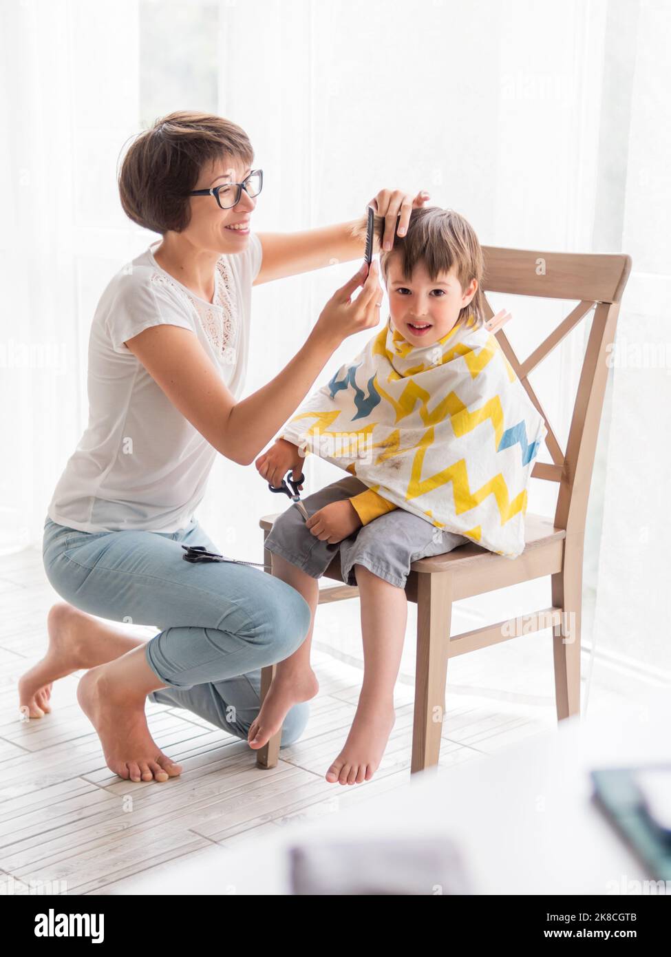 La madre taglia i capelli del figlio da sola. Il ragazzino si siede, coperto di stoffa, e tiene un paio di forbici. Vita familiare. Foto Stock