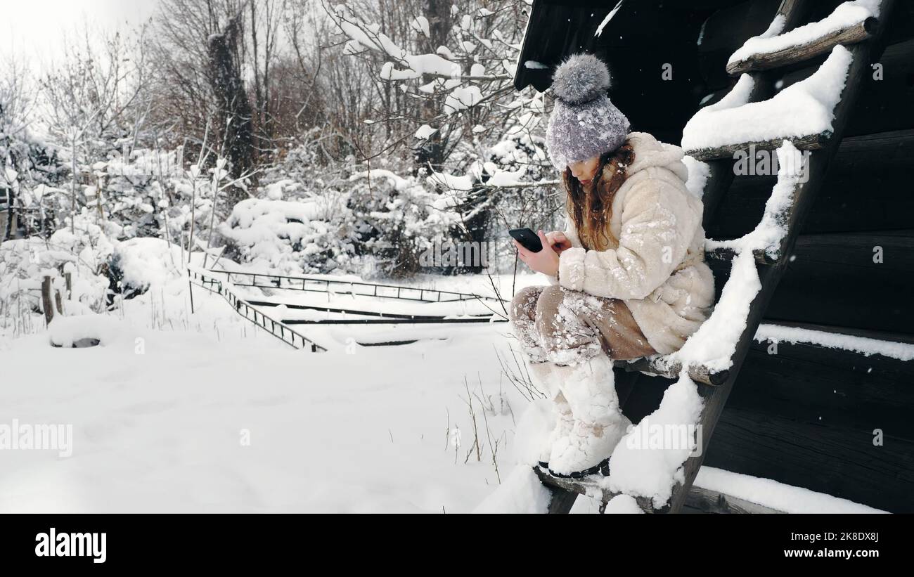 divertimento invernale innevato. la ragazza cutie, vestita con abiti invernali caldi, completamente coperta di fiocchi di neve, è seduta su panca e utilizza smartphone, all'aperto, durante le nevicate, vicino a una vecchia casa di legno. giornata invernale innevata. Foto di alta qualità Foto Stock