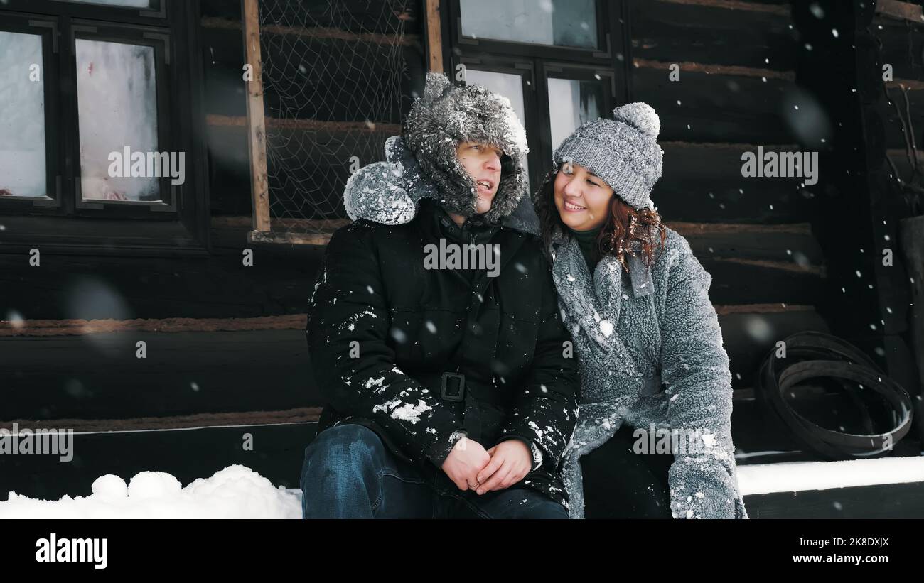 ritratto invernale. Felice, divertente coppia innamorata, uomo e donna, vestiti con abiti caldi d'inverno, completamente coperti di fiocchi di neve, sono seduti sulla panchina di una vecchia casa di legno e abbracciando, parlando. Buon tempo nella nevosa giornata invernale. Foto di alta qualità Foto Stock