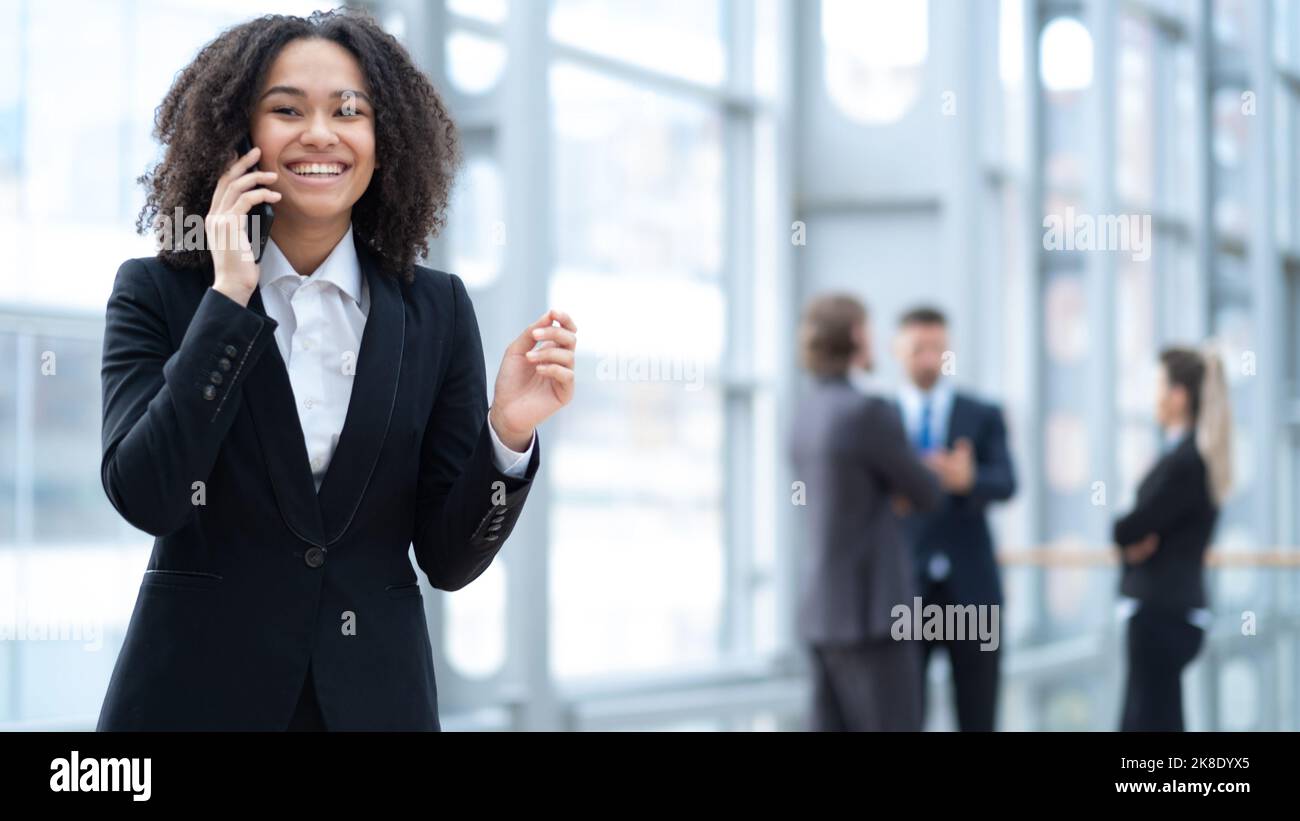Donna d'affari sicura che parla al telefono, leader femminile del team che si pone per la foto, in piedi in un ufficio moderno con team diversi sullo sfondo Foto Stock