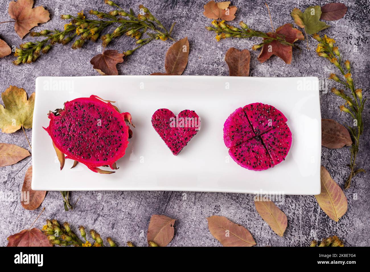 Piatto rettangolare con mezza pitahaya che espone l'interno rosa e con semi neri, pezzi pronti per mangiare, e un pezzo a forma di cuore al centro di Foto Stock