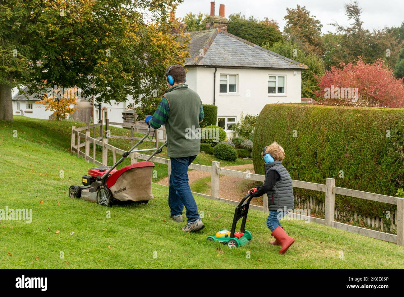Padre e figlio che falciano l'erba, il ragazzo carino che spinge un rasaerba giocattolo indossando cuffie che imitano e seguono il suo papà, Regno Unito Foto Stock