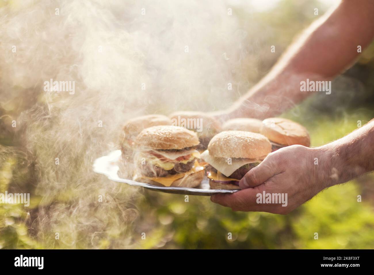 Mano dell'uomo che serve cheeseburgers in mezzo al fumo nel cortile Foto Stock