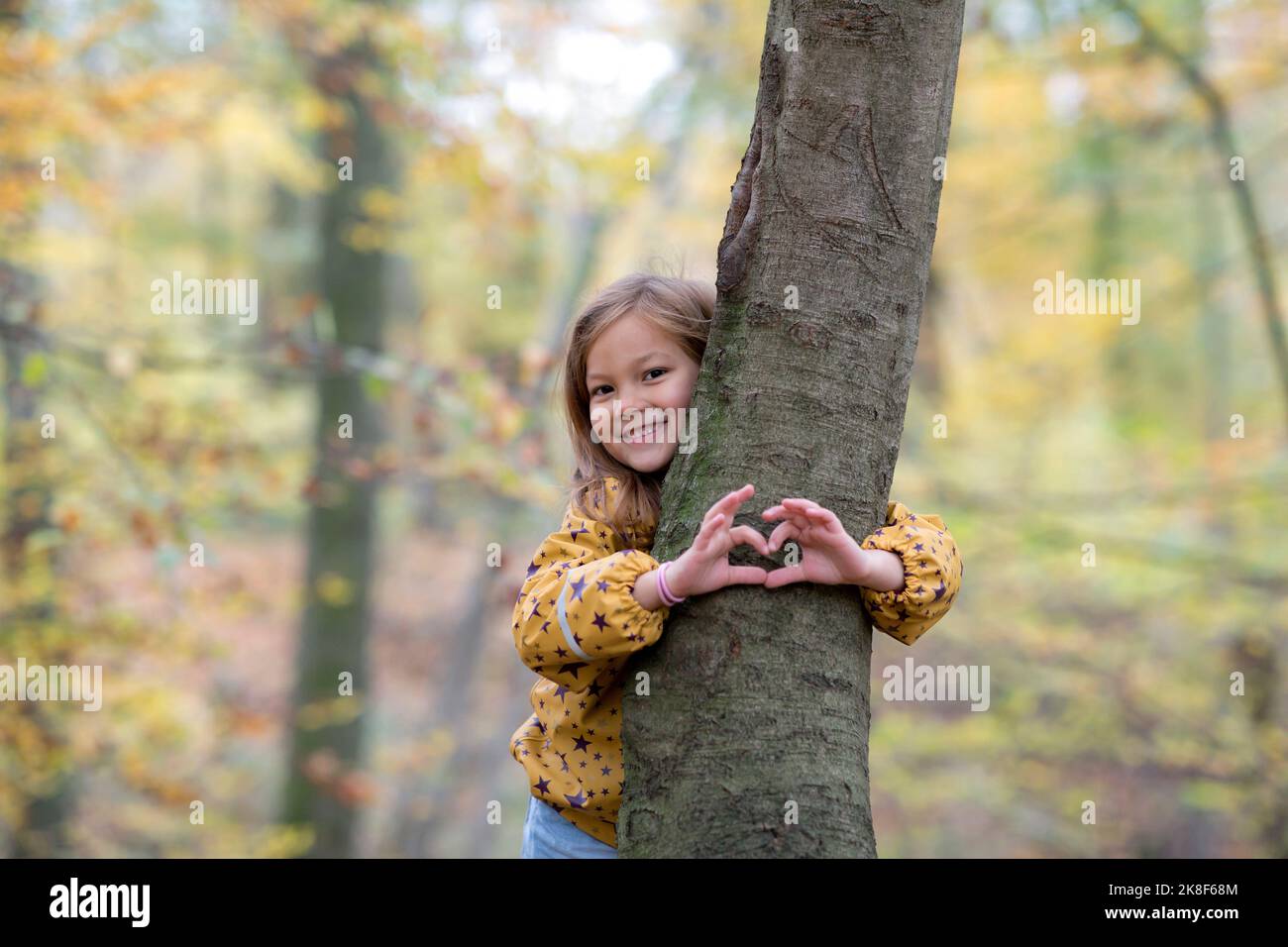 Sorridente ragazza carina che abbraccia l'albero e la forma del cuore gesturante nella foresta Foto Stock