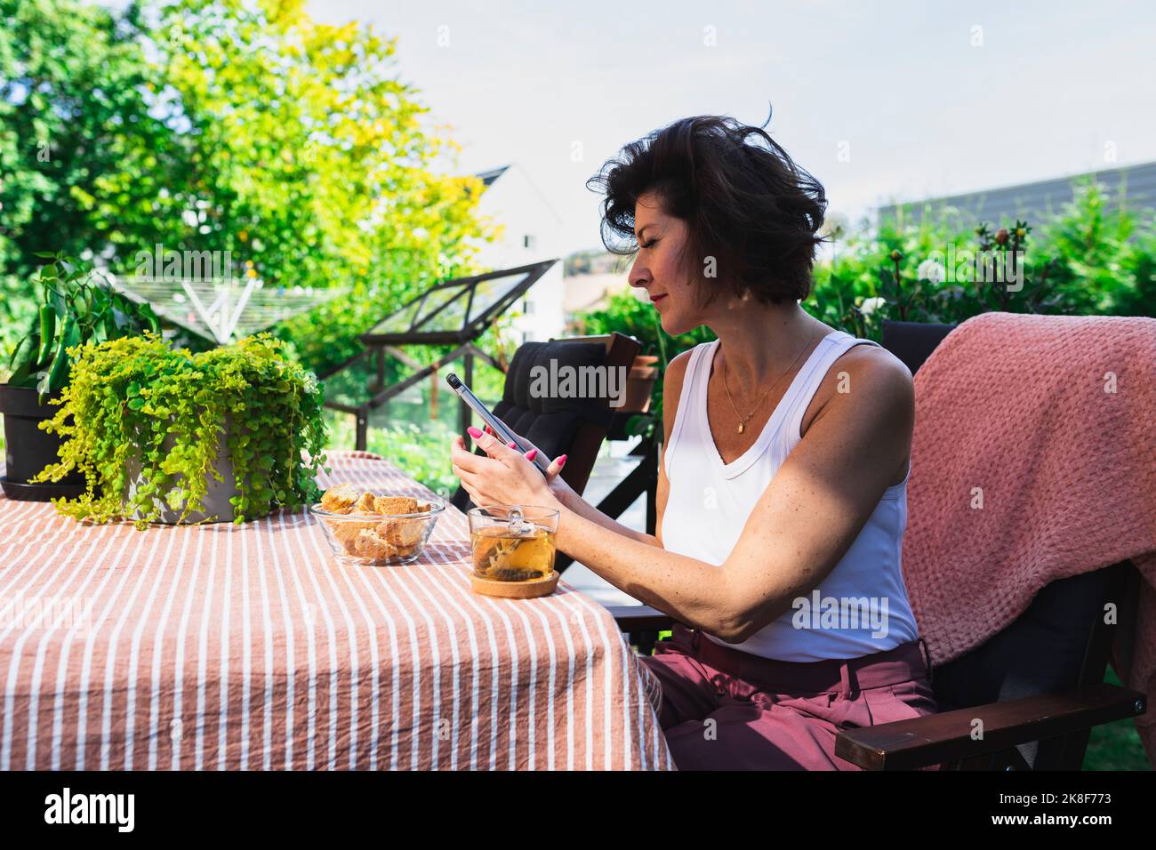 Donna matura con snack e tea Cup sul tavolo in terrazza con telefono cellulare Foto Stock