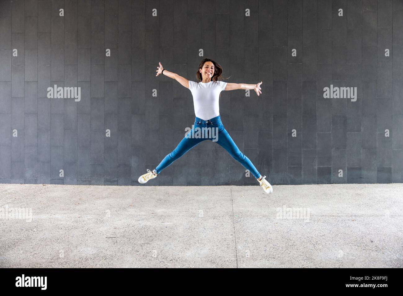 Donna felice con braccia allungate saltando sul sentiero davanti alla parete grigia Foto Stock