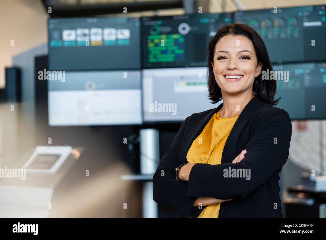 Donna d'affari sorridente con le braccia incrociate davanti ai monitor dei computer Foto Stock