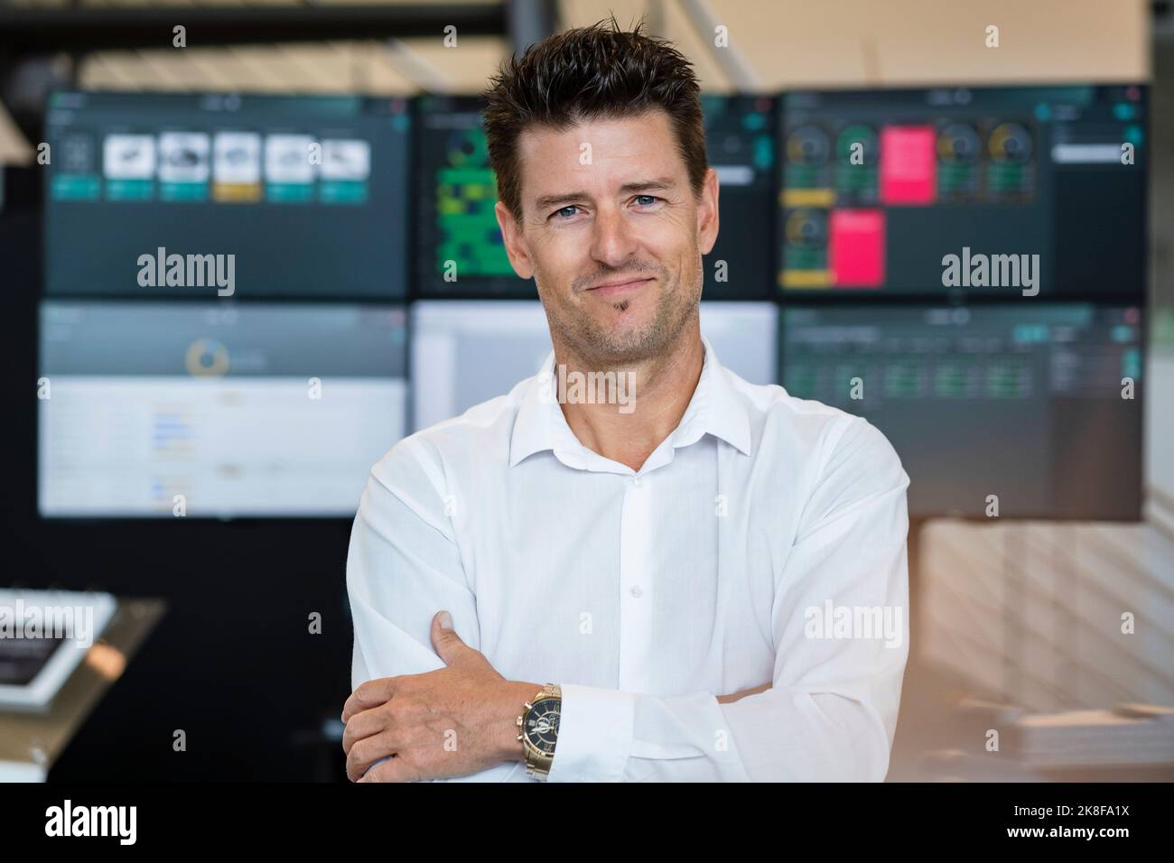 Uomo d'affari sorridente con le braccia incrociate davanti ai monitor dei computer Foto Stock