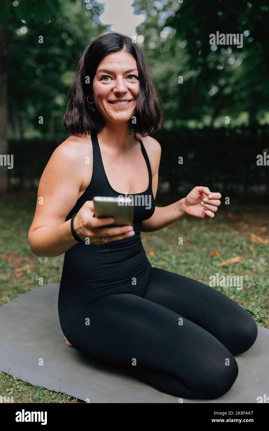 Sorridente insegnante di yoga con telefono cellulare seduto sul tappetino per esercizi nel parco Foto Stock