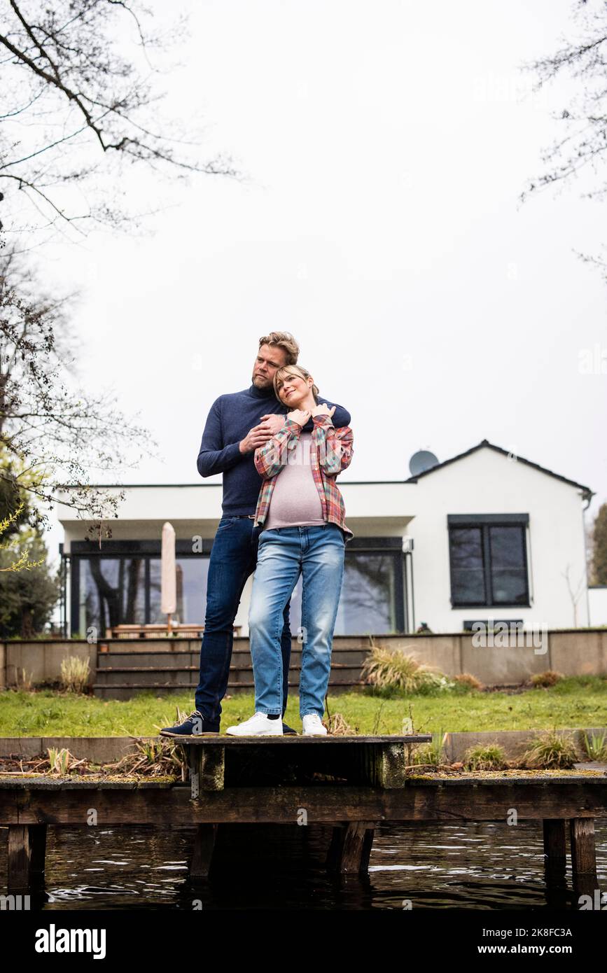 Uomo che abbraccia donna incinta da dietro sul molo di fronte a casa Foto Stock