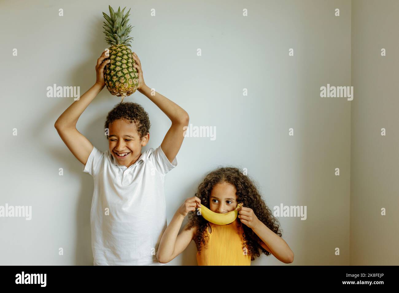 Ragazzo felice che tiene l'ananas sopra la testa da sorella coprendo le labbra con la banana di fronte al muro Foto Stock