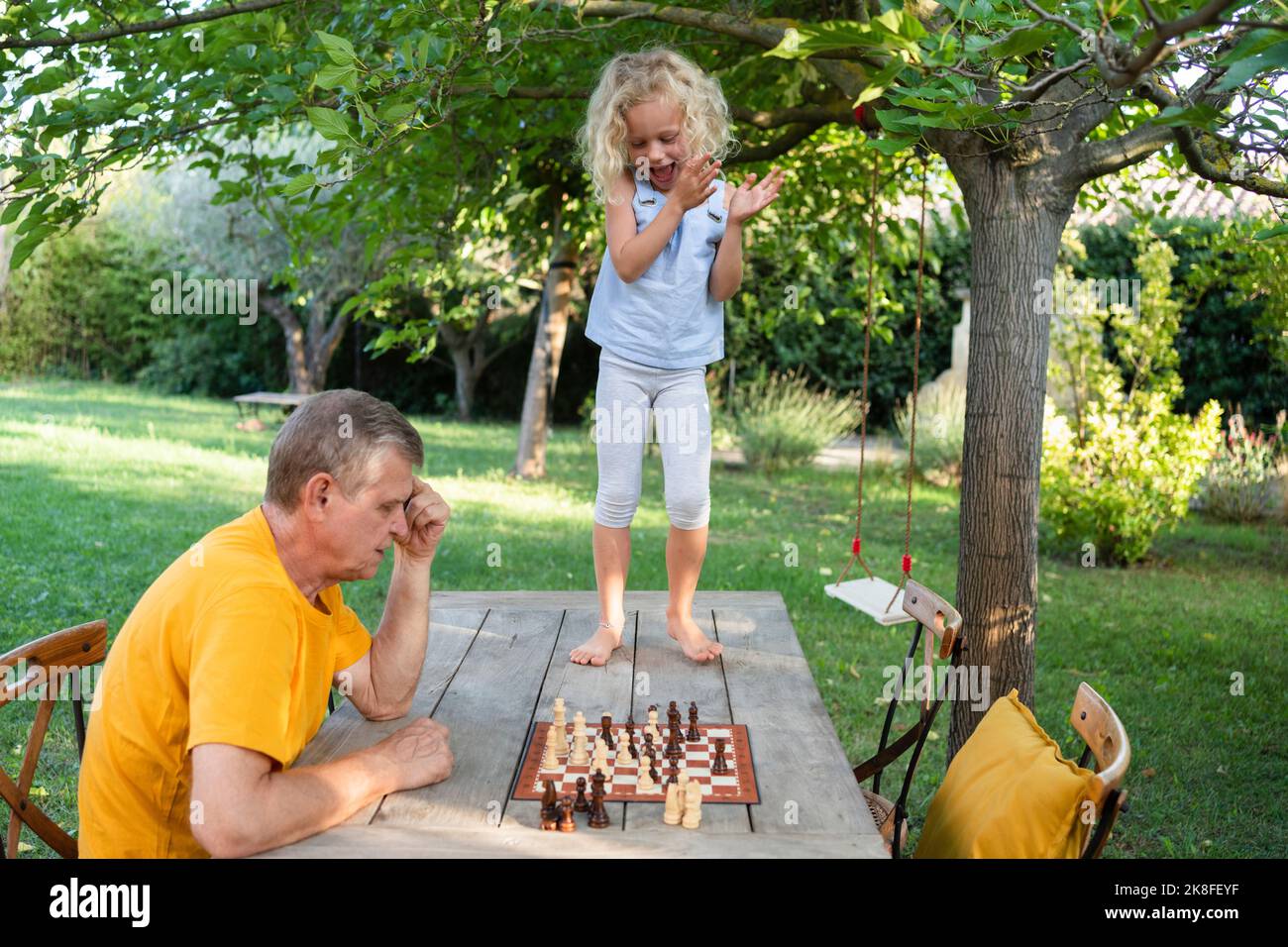 Ragazza carina che aggrappa le mani da nonno giocando a scacchi sul tavolo in giardino Foto Stock