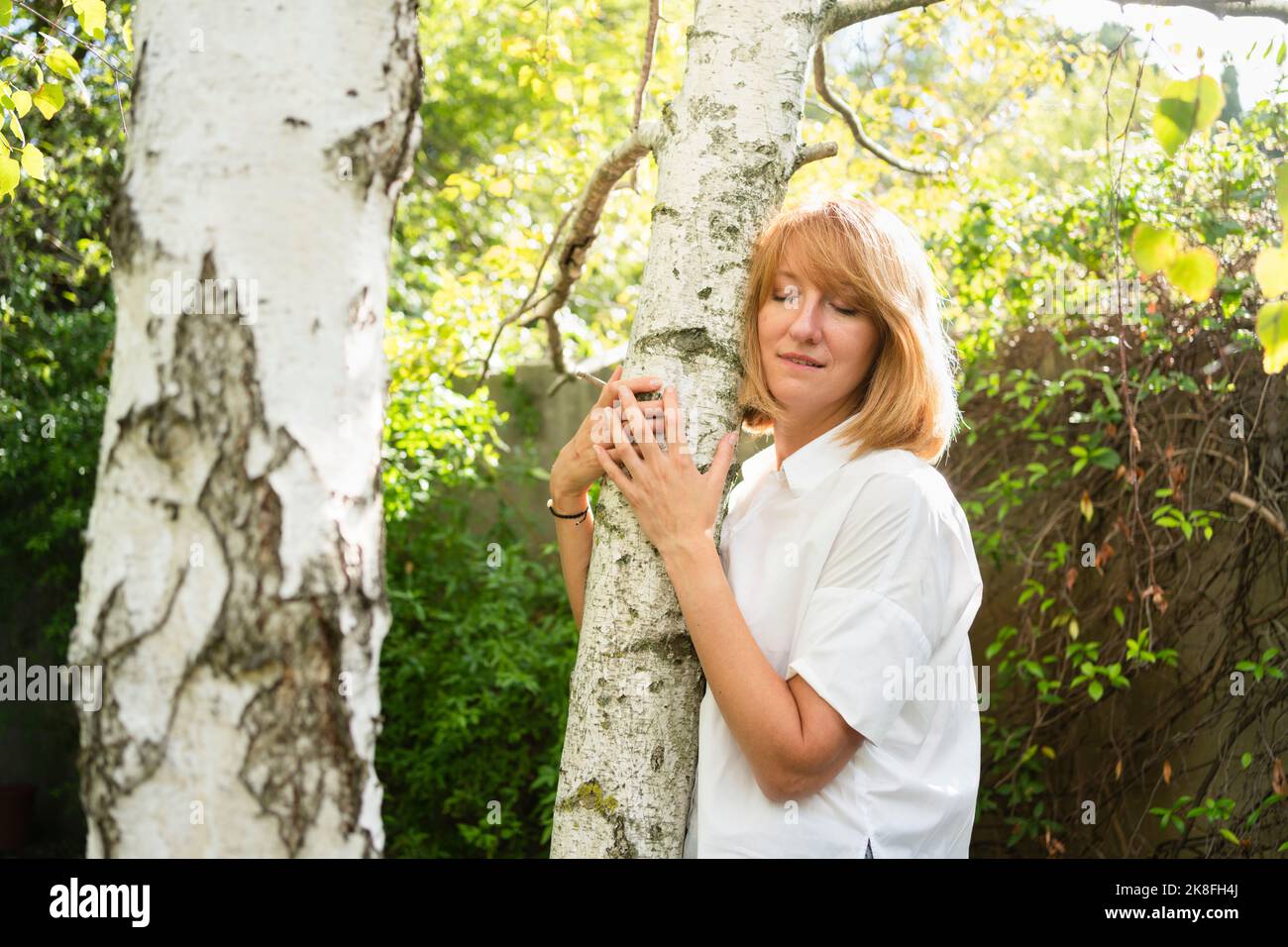 Donna matura con gli occhi chiusi abbracciando tronco di albero in giardino Foto Stock
