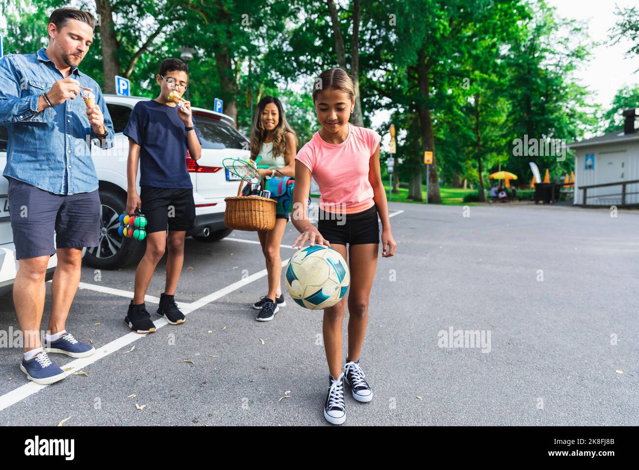 Ragazza che gioca con la palla da parte della famiglia godendo gelato al parcheggio Foto Stock