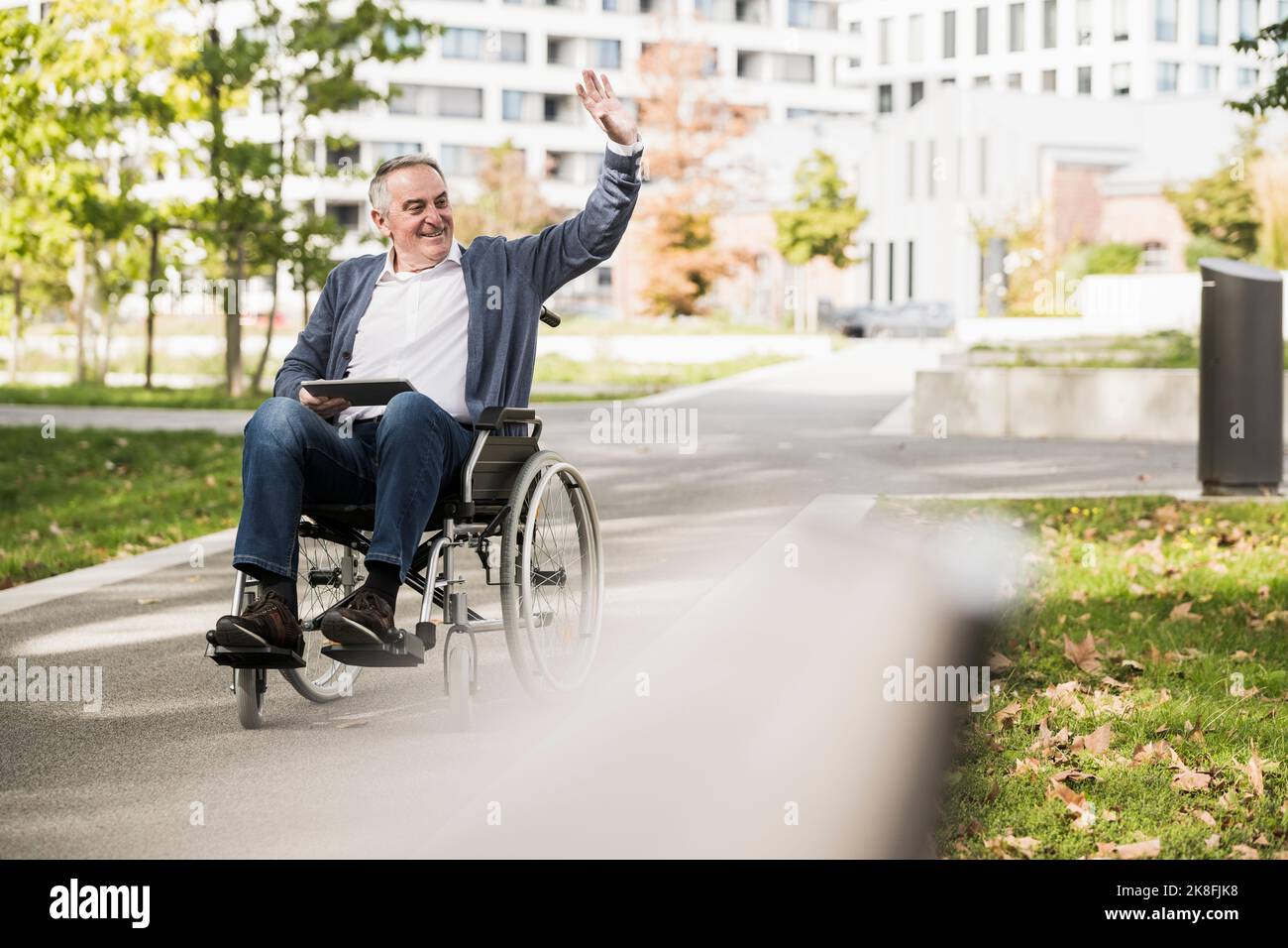 Uomo anziano sorridente con un tablet che ondilla e si siede su sedia a rotelle Foto Stock