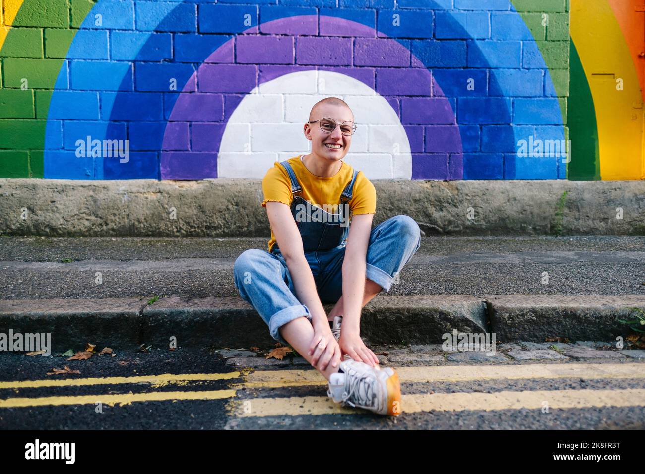 Felice transgender persona seduta sul sentiero di fronte al muro arcobaleno Foto Stock