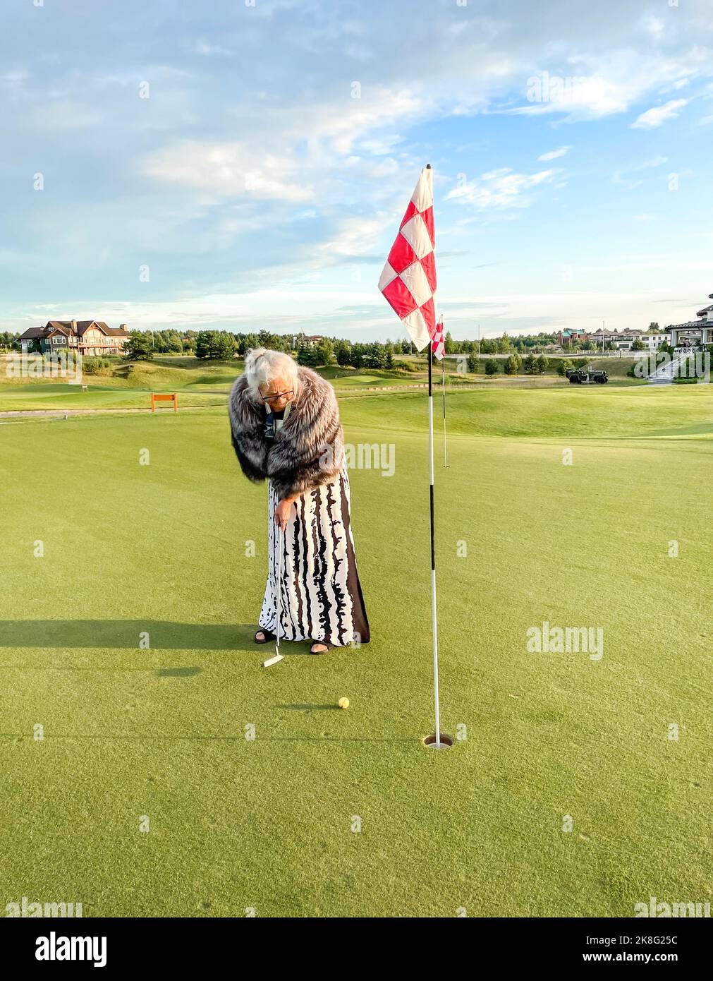Donna anziana gioca a golf sul campo da golf. Segna la palla nel buco con la bandiera a scacchi rossa e bianca. Foto Stock