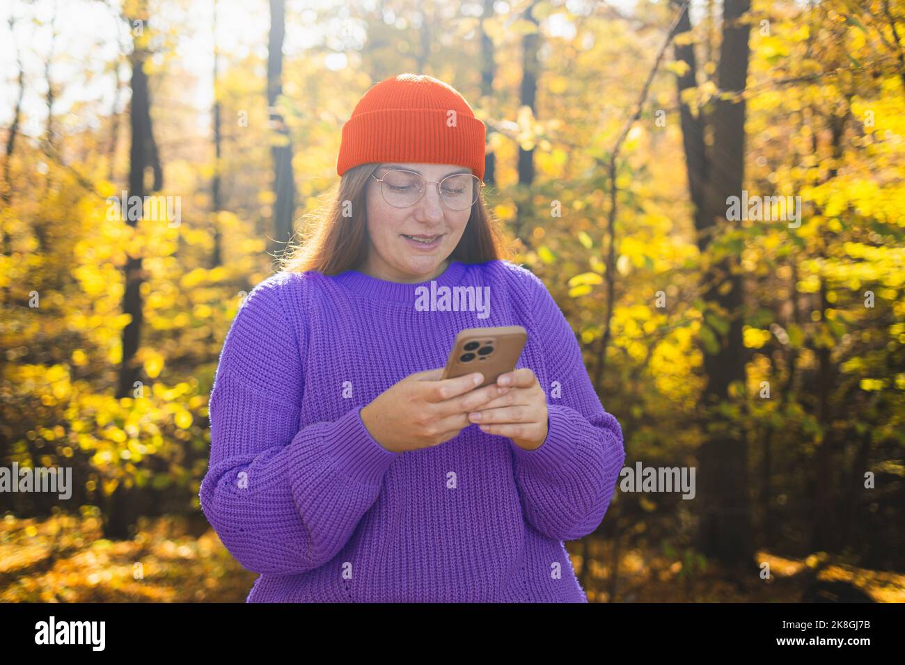 Allegro giovane ragazza bella e gioiosa che indossa abiti eleganti camminando nella foresta autunnale, guardando il telefono cellulare Foto Stock