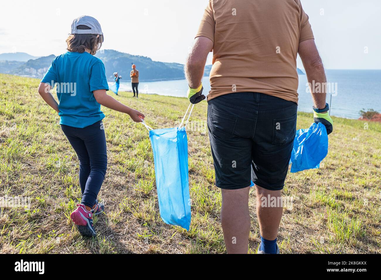 Eco attivisti di diverse età in abbigliamento sportivo con sacchi di bidone blu raccolta rifiuti su verde collina erbosa vicino al mare blu calmo nelle giornate di sole Foto Stock